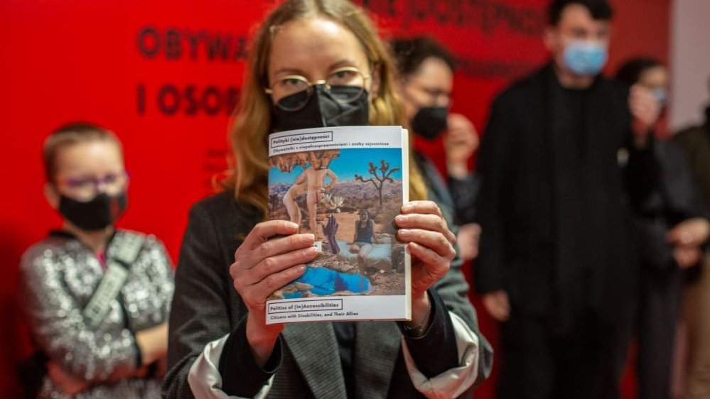 Photo from the exhibition: a woman is showing an exhibition catalogue.