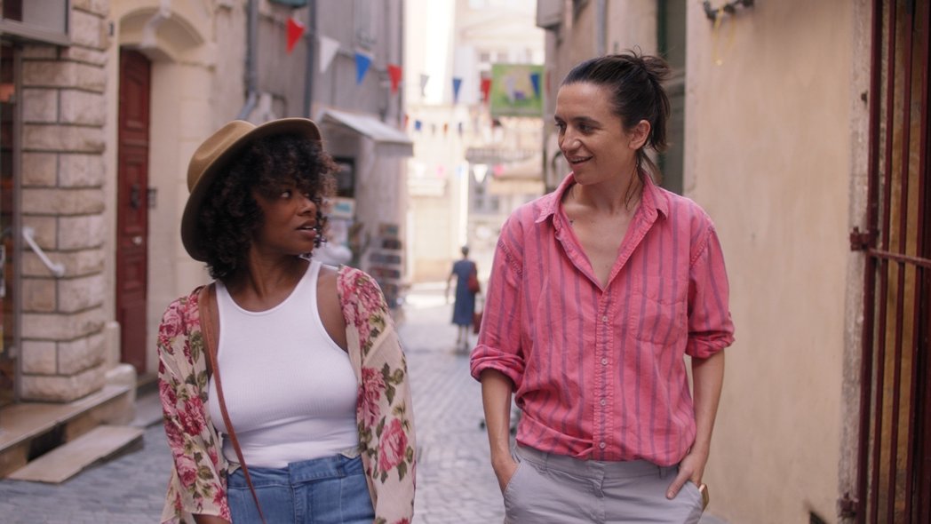 The photo shows two women walking down the street on a sunny day looking at each other.