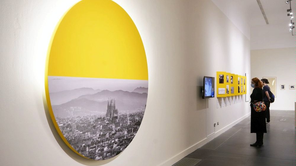 Photo of the exhibition hall with the exhibits from the exhibition. Pictures and a monitor hanging on the walls and a few people watching the exhibition. - grafika artykułu