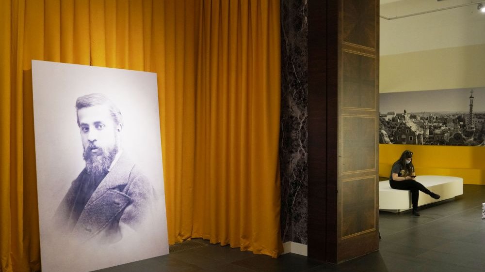 One of the exhibition rooms with Gaudi's portrait leaning against the wall. In the background, another room and a person sitting on a seat.