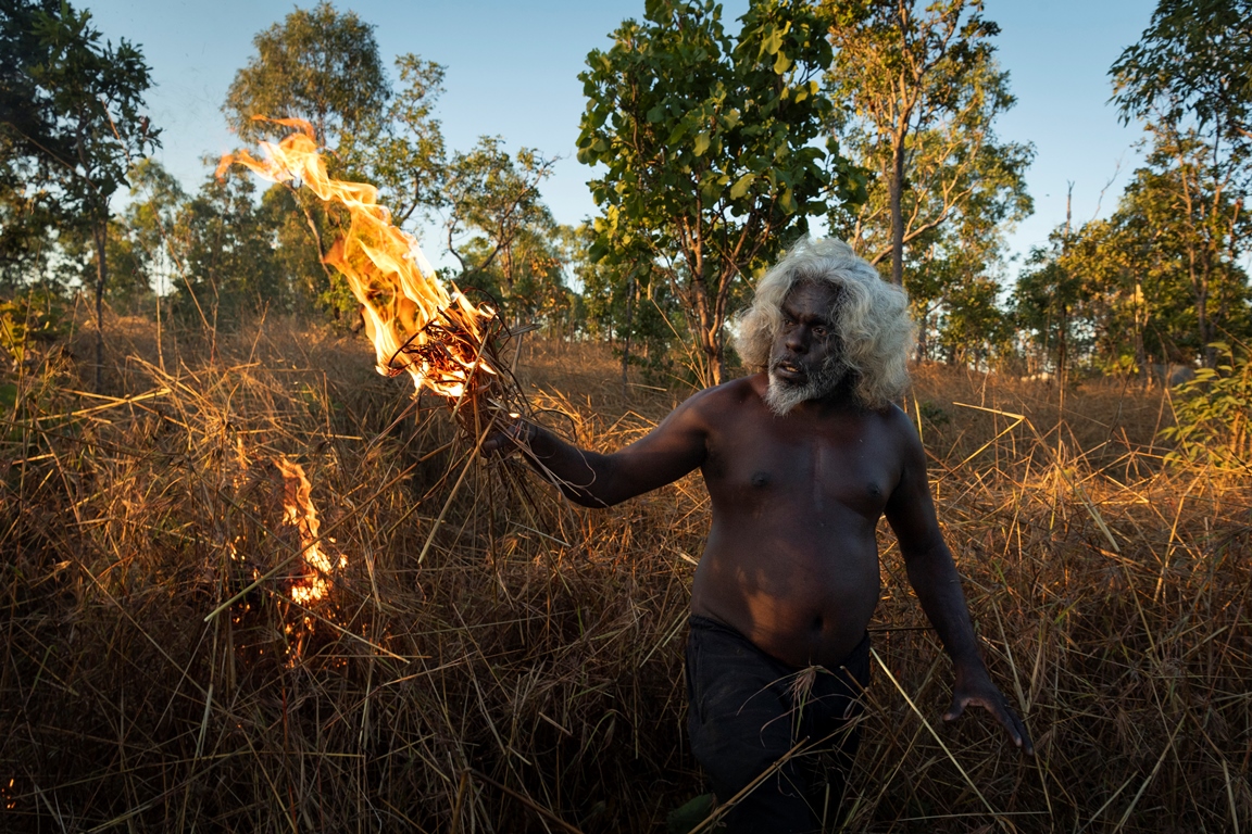 Photo from the exhibition: the indigenous Australian holding some dry grass, which is burning. Some trees in the background - grafika artykułu