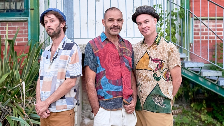 Photo of the band: three men in colourful shirts; as a background - a part of a building with stairs and some plants.