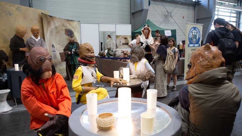Four people in disguise sitting at a table. Several people in disguise in the background.