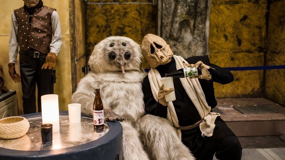 Two people in disguise sitting at a table, one of them is pouring a beverage from a bottle to a cup.