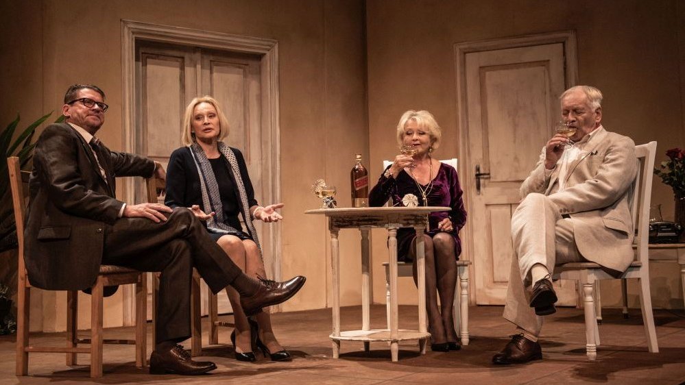 A scene from the performance - four actors sitting at a table, two of them drinking an alcohol from glasses.