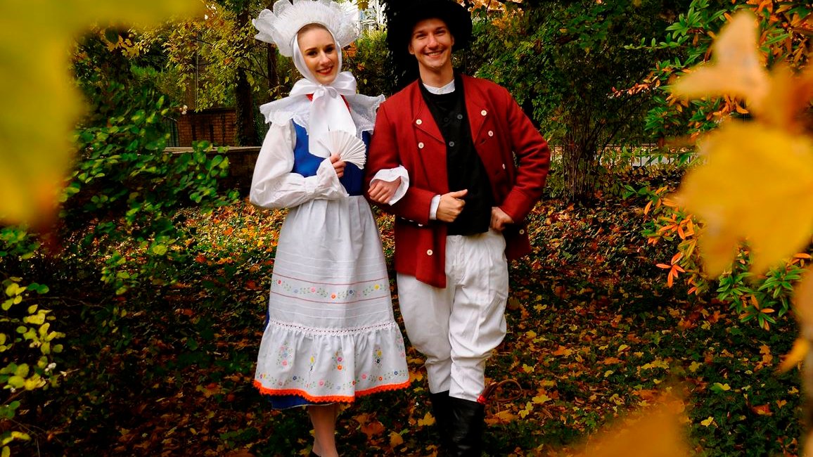 Photo of a woman and a man dressed in traditional folk costumes. Trees and colourful leaves as a background.