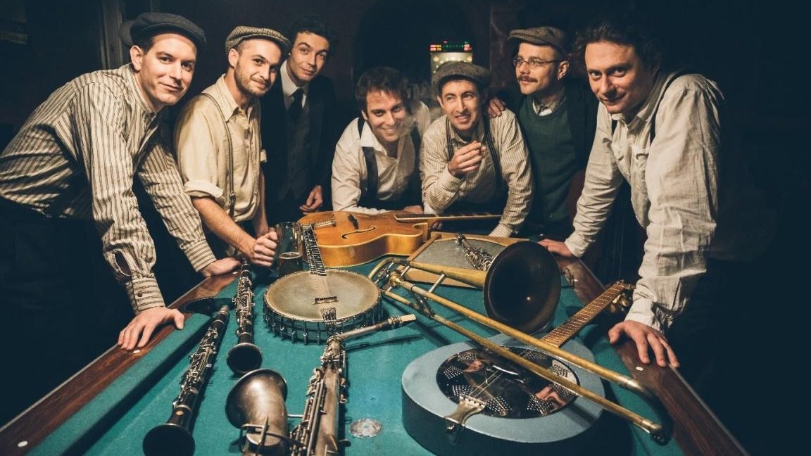 Photo of the band - seven men standing at the billiard table, on which various musical instruments are placed.