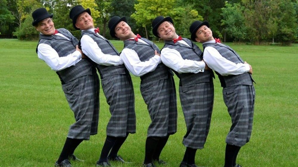 Photo of the band - five men standing in a row on a meadow. Trees as a background.