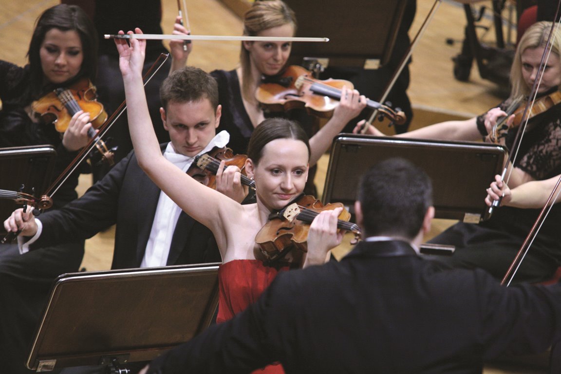 Photo of Agata Szymczewska playing the violin on stage. The conductor in front of Agata Szymczewska and the musicians of the orchestra behind her. - grafika artykułu