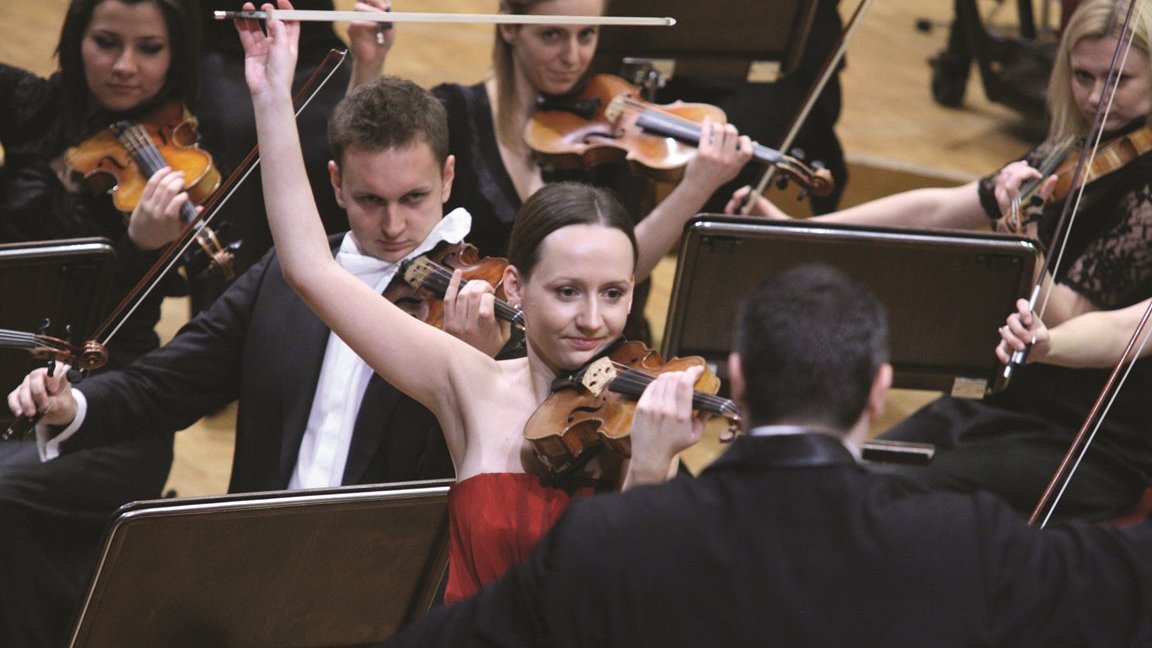 Photo of Agata Szymczewska playing the violin on stage. The conductor in front of Agata Szymczewska and the musicians of the orchestra behind her.