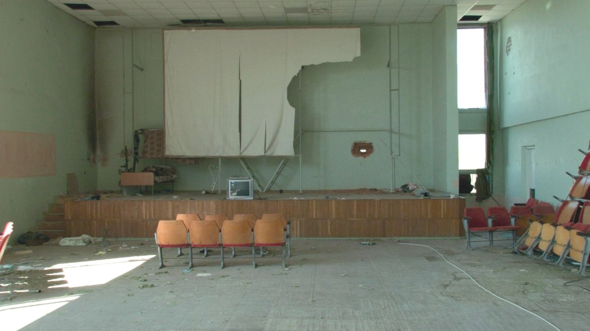 The interior of a hall with destroyed screen on the wall. Under the screen there is a stage with a small TV sat on it. In front of the stage 8 chairs. The whole hall is messy. - grafika artykułu