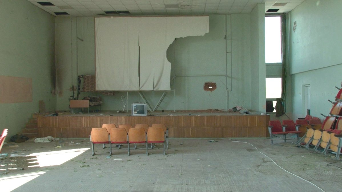 The interior of a hall with destroyed screen on the wall. Under the screen there is a stage with a small TV sat on it. In front of the stage 8 chairs. The whole hall is messy.