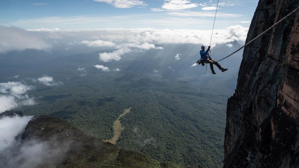 Photo of a man hanging on a rope by the rock. Clouds and mountains covered with forest as a background.