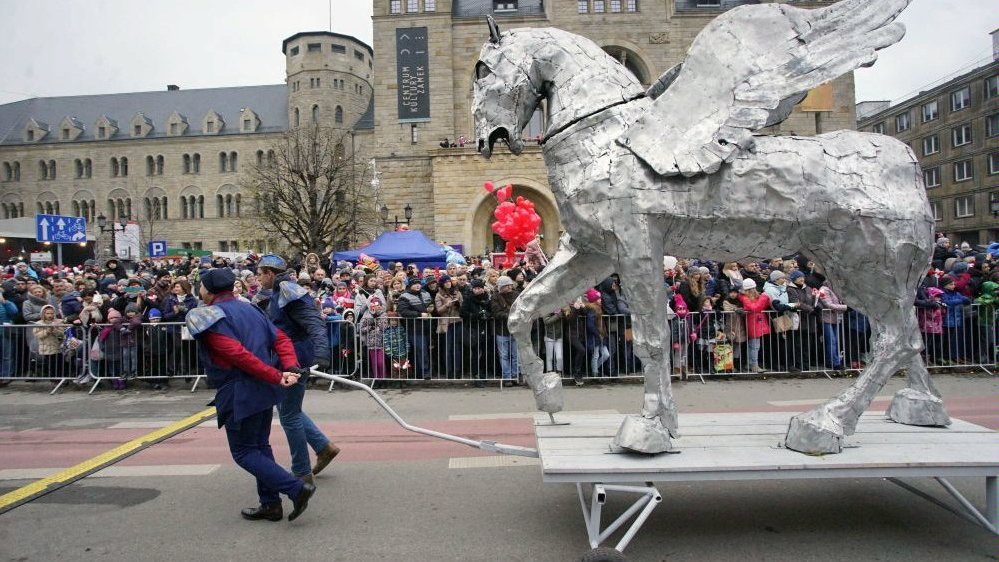 Photo from one of the previous St. Martin parade: picture of St. Martin street with performers (two men pulling a big statue of silver Pegasus on a platform) and people watching the show. The Castle Culture Centre in the background.