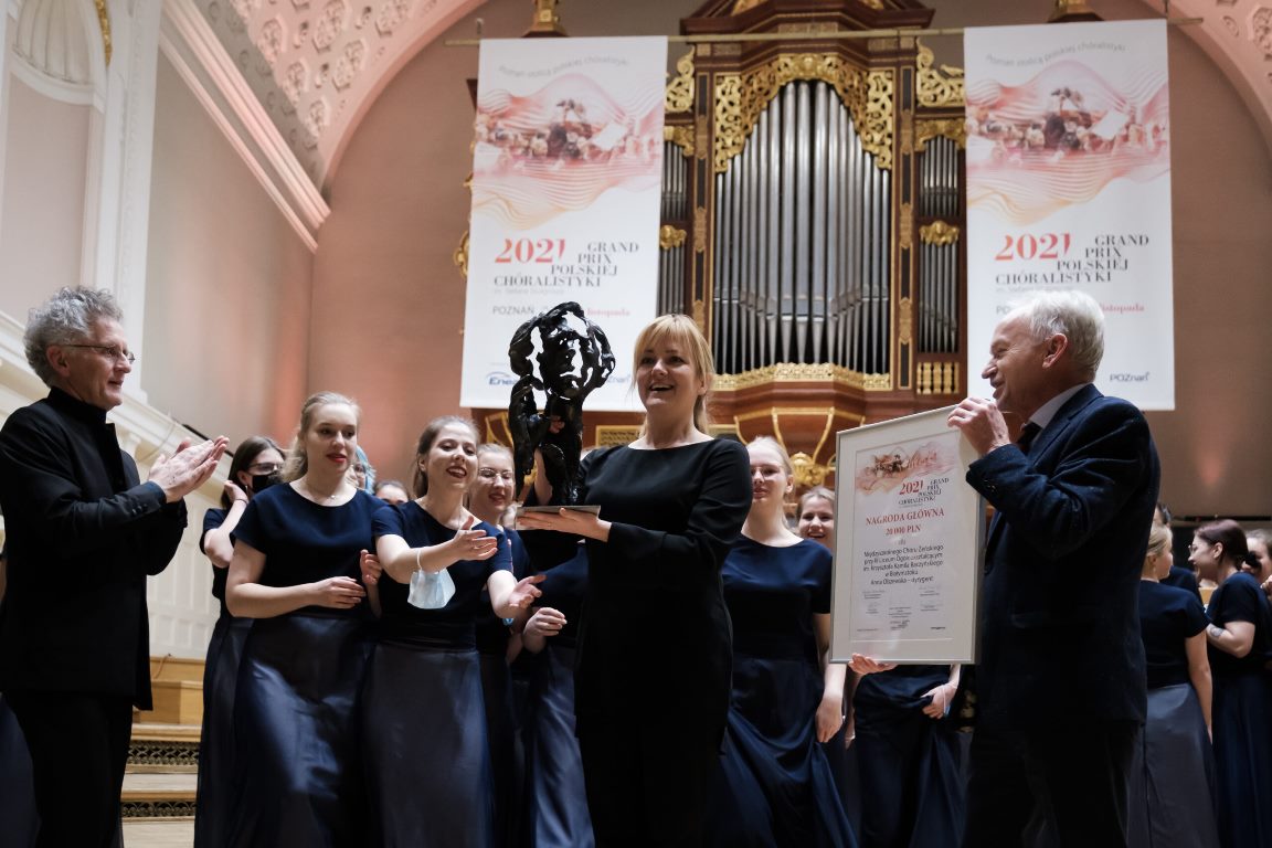 Picture of the winners - the choir's woman conductor holding a statuette and two men - one of them holding a diploma and the other clapping his hands. Behind the conductor girls members of the choir. Pipe organ in the background. - grafika artykułu