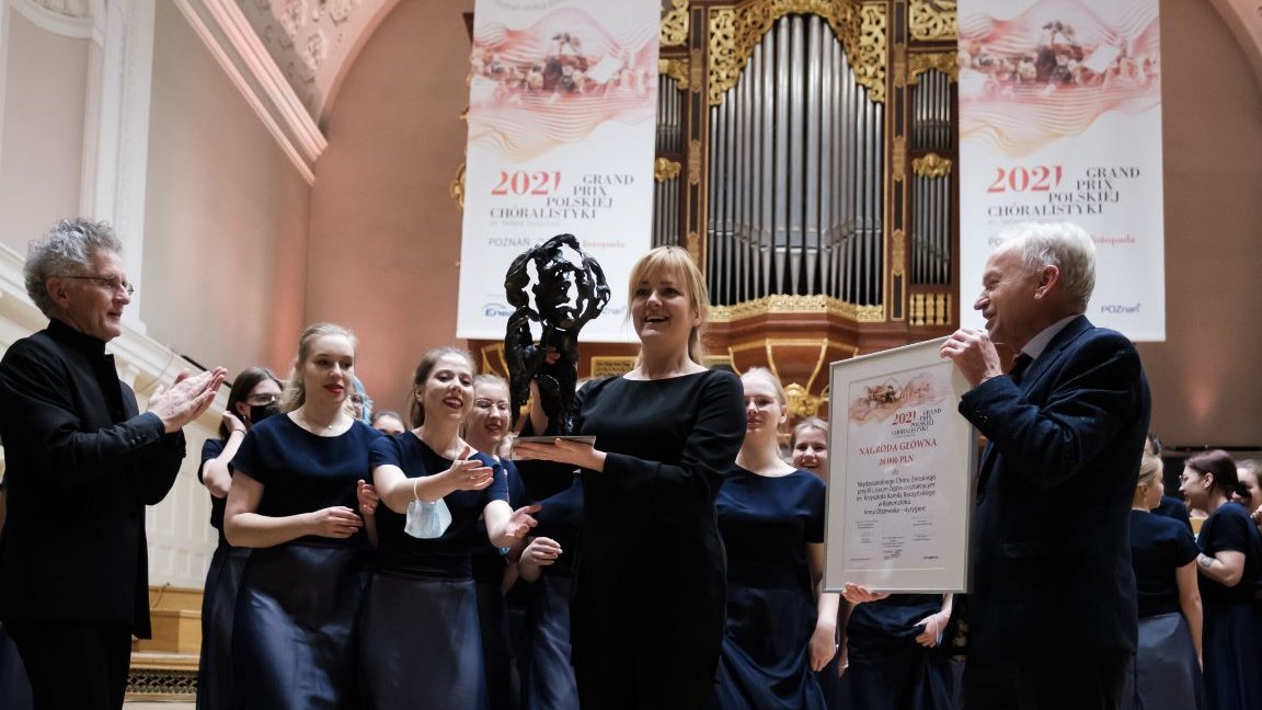 Picture of the winners - the choir's woman conductor holding a statuette and two men - one of them holding a diploma and the other clapping his hands. Behind the conductor girls members of the choir. Pipe organ in the background. - grafika artykułu
