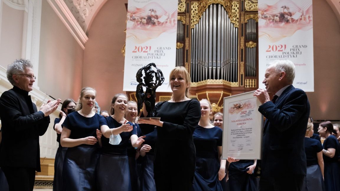 Picture of the winners - the choir's woman conductor holding a statuette and two men - one of them holding a diploma and the other clapping his hands. Behind the conductor girls members of the choir. Pipe organ in the background.