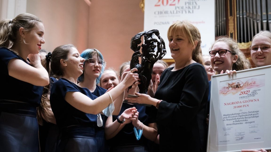 Photo of a woman conductor holding a statuette, girl members of the choir around her, one of them holding a diploma.