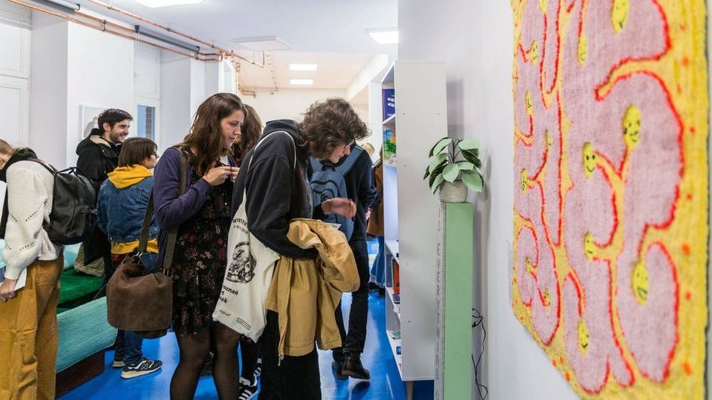 Photo of people watching the exhibition. In the foreground one of an artist's works hanging on the wall.