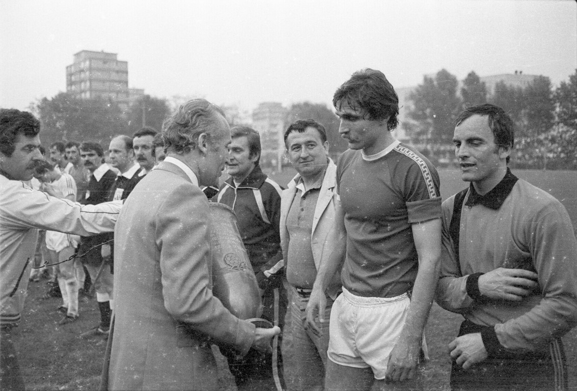 Black and white photo of men standing in a row. Opposite one of them a man holding a cup. In the background grass, trees and buildings. - grafika artykułu