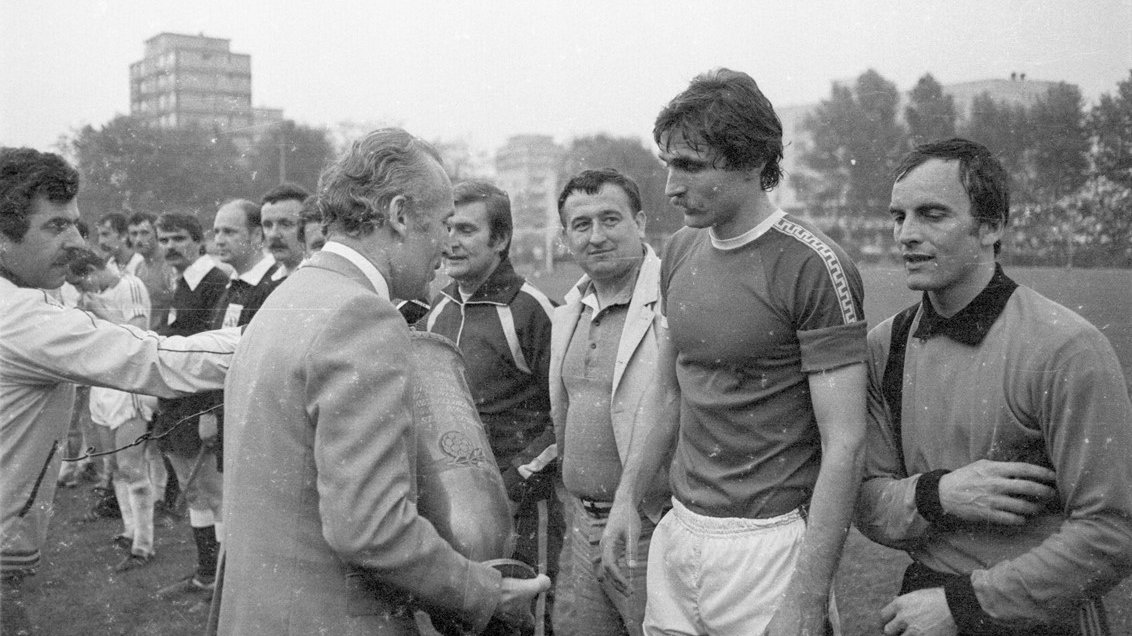 Black and white photo of men standing in a row. Opposite one of them a man holding a cup. In the background grass, trees and buildings.