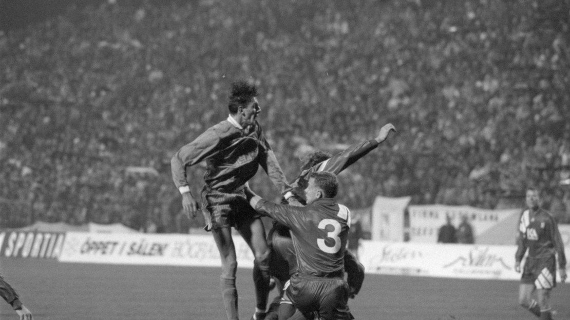 Black and white photo of three soccer players on the football field, fighting for the ball. A stadium full of fans in the background.