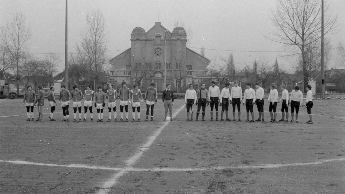 Black and white photo of two teams standing in a row on a football field. Building and trees in the background.