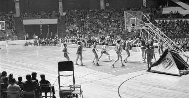 Black and white photo of a basketball game in a sports hall. Tribunes with fans around the pitch.