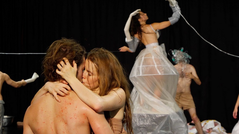 Photo from the performance. In the foreground a kissing couple. In the background a woman in a white transparent dress. - grafika artykułu