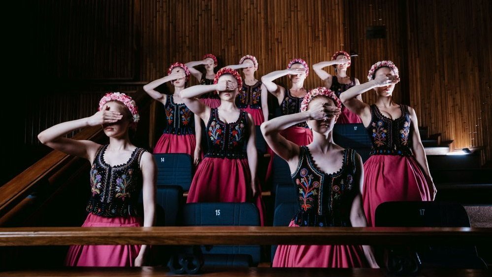 Photo from the performance: women in folk costumes, standing in four rows; each of them covers her eyes with her right hand.