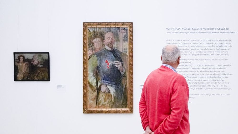Photo of an exhibition hall with two paintings and a text written on the wall. A man is reading a text on the wall.
