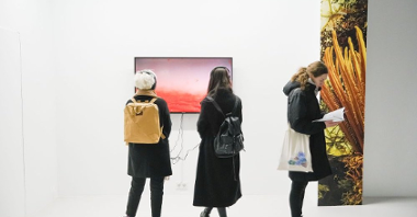 Exhibition hall: two people watching the picture displayed on a TV screen and one person reading a book.