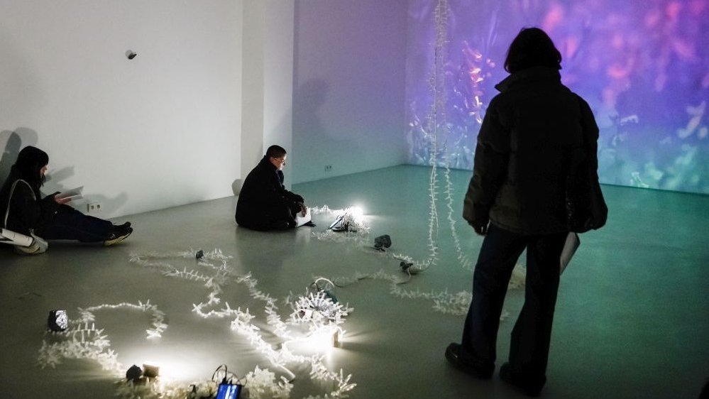 Exhibition hall: three people in it and a picture displayed on a wall and a few lightening items on a floor.