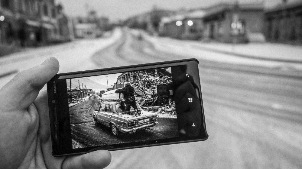 A black and white photo of a hand holding a mobile phone, in which there is a picture of a road with a car and a man on it. In the background the same empty road as in the picture.