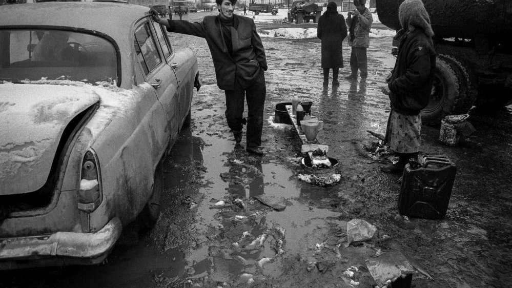Black and white photo of an old car on a muddy road. A man leaning against a car and looking at the woman, who stands opposite him.