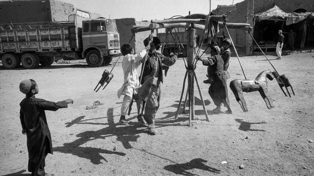 Black and white photo of a wooden merry-go-round and a few people by it. In the background a lorry and some buildings.
