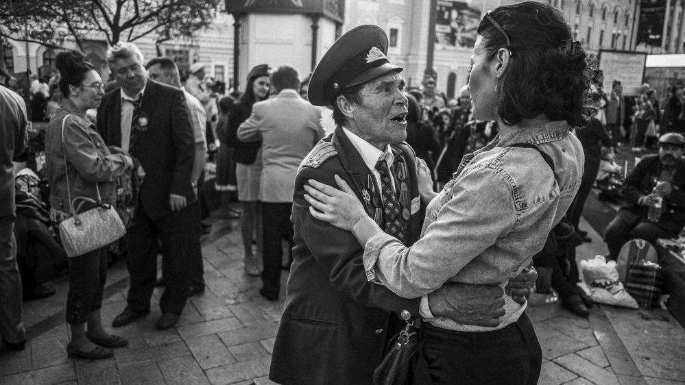 Black and white photo of an older man in a military uniform, who is holding a woman in his arms. In the background other people and buildings.