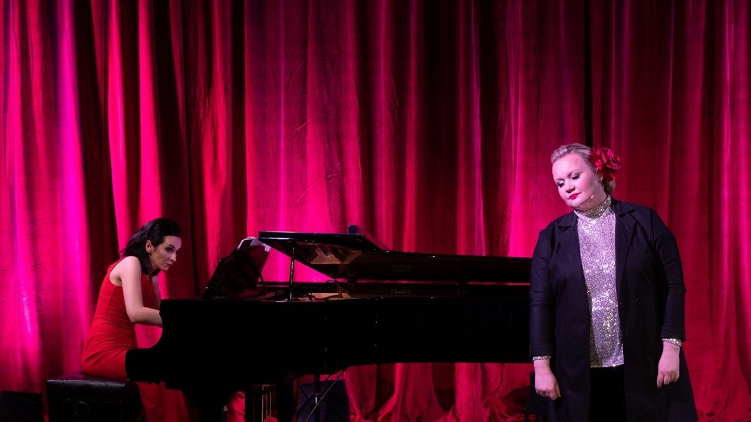 Photo of two women: one is standing next to the piano, another is playing the piano.