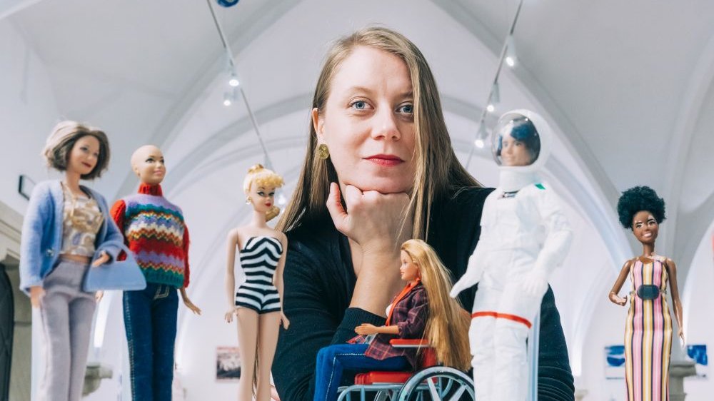 Photo of Aleksandra Podżorska, curator of the exhibition. In front of her four Barbie dolls, one of them on a wheelchair. - grafika artykułu