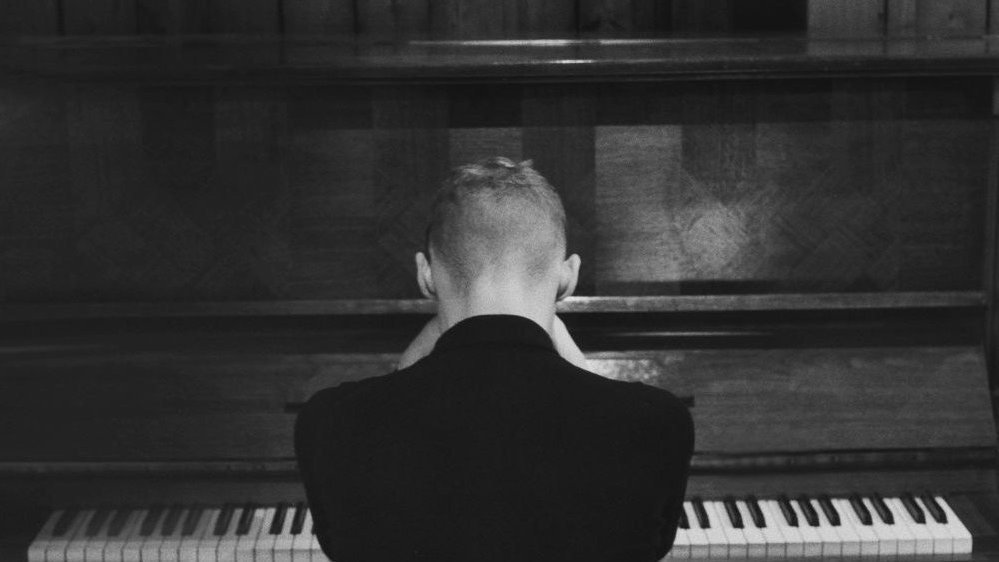 Black and white photo of Igor Herbut, who sits at the piano back to the camera. - grafika artykułu