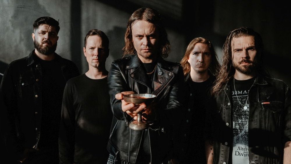 Photo of the band - five men in black clothes, one of them is holding a copper chalice in his hand.