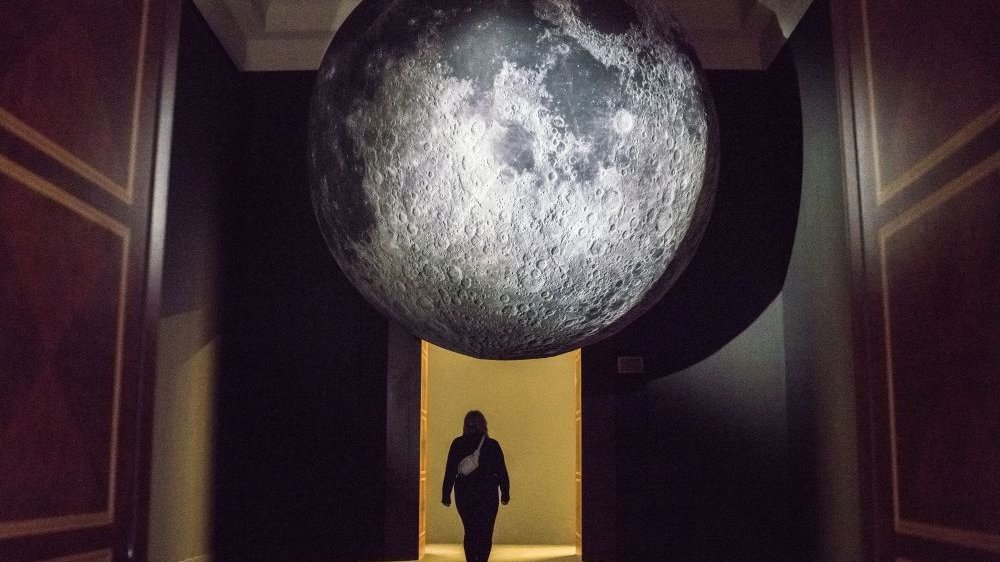 Photo of a dark room, on the end of which there is a door. Under the ceiling a model of a silver planet and under it a woman crossing towards the door.