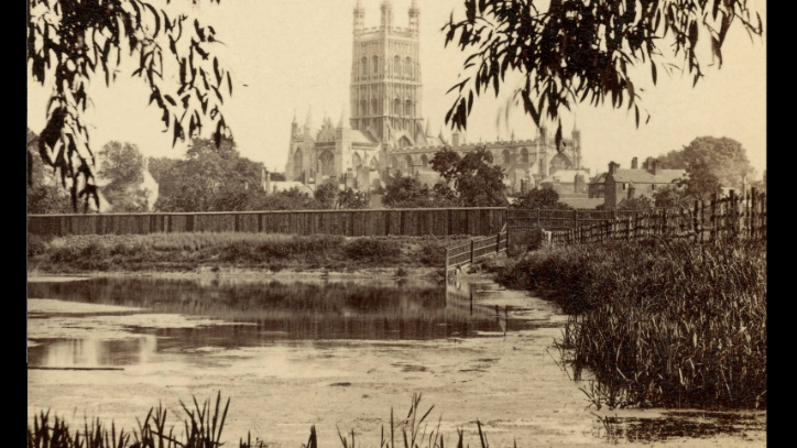 Sepia photograph of the Gloucester Cathedral, which is in the background. In the foreground a lake or a river with some water plants.