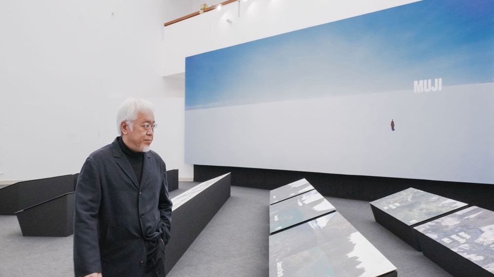 Photo of Kenya Hara standing by his works displayed on low expositors. In the background a big picture in blue and white colours with inscription "Muji".