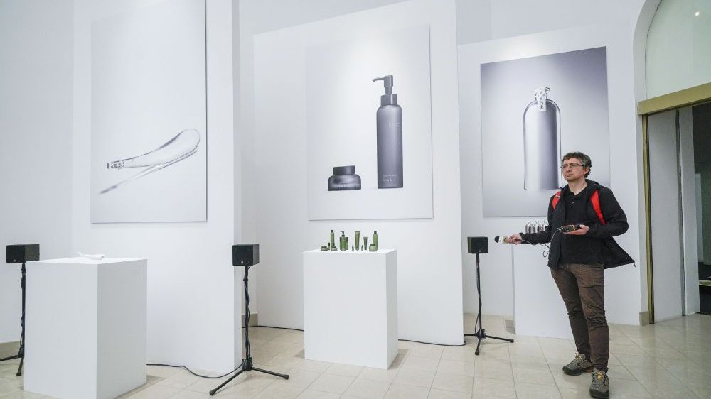 Photo of the exhibition hall with three pictures hanging on the walls. Under the pictures white expositors with exhibits. On the right a man standing and looking straight ahead.