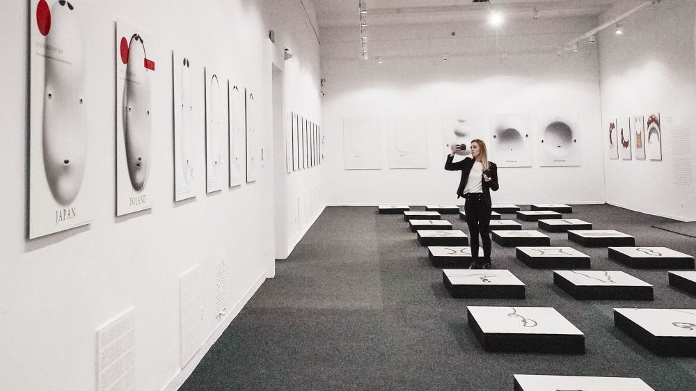 Photo of the exhibition hall with pictures displayed on the floor and walls. One woman looking at pictures on the wall.