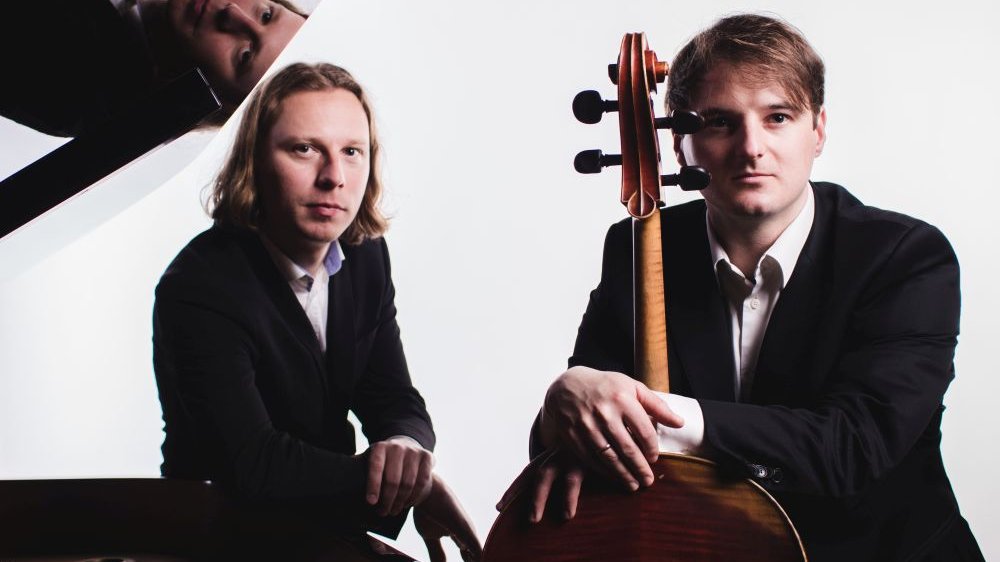 Photo of FudalaRot Duo - two men stending by their musical instruments: the piano and the cello.