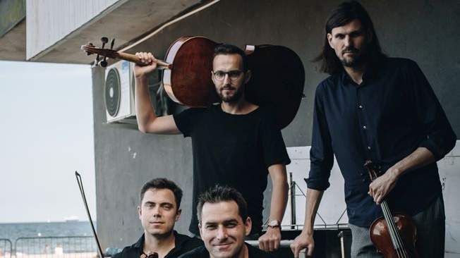 Photo of Atom String Quartet - a four-men band, two of them sitting, another two standing; each of them is holding a musical instrument. A wall of a building and the sea-side as a background.