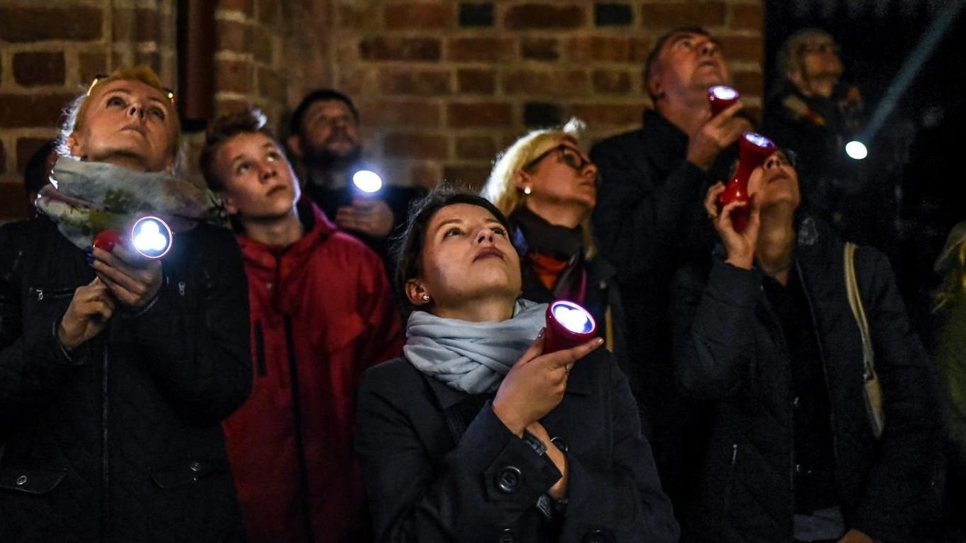 Photo of a group of people with torches, looking up. The brick wall as a background.