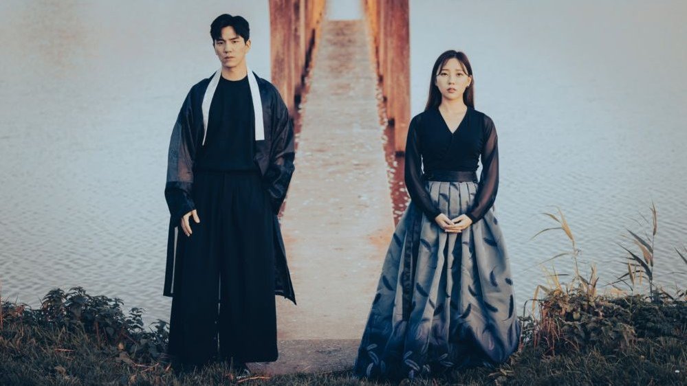 Photo of a young woman and a young man standing on a bank of the river. Behind them a narrow bridge.